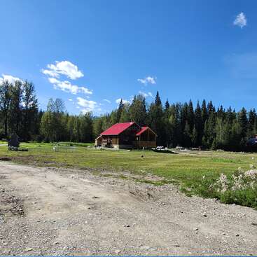 A cozy cabin with a red roof sits in the middle of a grassy clearing, surrounded by dense forest under a bright blue, cloud-dotted sky.