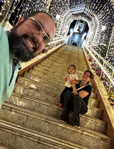 A man takes a selfie with a smiling woman and child sitting on decorated stairs, illuminated by festive string lights leading up to a church at night.
