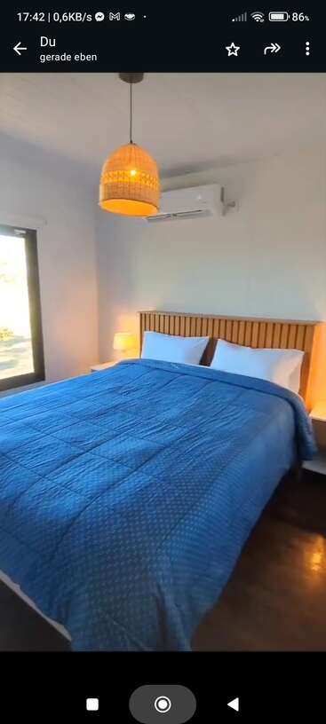 Cette image montre une chambre à coucher confortable avec un grand lit recouvert d'une couverture bleue, des oreillers blancs, un éclairage doux, une tête de lit en bois, une fenêtre, une table de nuit et un climatiseur.