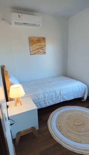 Une petite chambre confortable est équipée d'un lit simple avec une couverture blanche texturée, d'un tapis rond, d'une lampe de chevet, d'un tableau mural et d'un climatiseur.