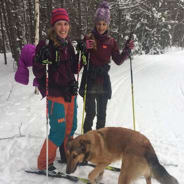 Deux femmes en vêtements d'hiver font du ski de fond dans une forêt enneigée, en souriant. Elles sont accompagnées d'un chien brun et profitent ensemble de l'aventure en plein air.