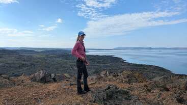 Une femme se tient sur un terrain rocheux surplombant un vaste paysage accidenté avec une grande étendue d'eau, sous un ciel bleu parsemé de nuages épars.
