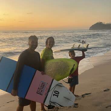 Trois personnes tenant des planches de surf se tiennent sur une plage de sable au coucher du soleil, souriantes. De douces vagues déferlent et une petite île apparaît au loin. Un moment paisible.