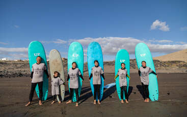 Seis pessoas estão em uma praia de areia, cada uma segurando uma prancha de surfe na posição vertical. Elas usam roupas de mergulho e camisas cinza combinando, sorrindo sob um céu azul claro.