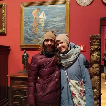 Das Bild zeigt einen Mann und eine Frau, die vor einer roten Wand stehen, die mit Kunstwerken geschmückt ist, darunter ein Gemälde mit einer Person auf dem Wasser und eine Skulptur einer Person.