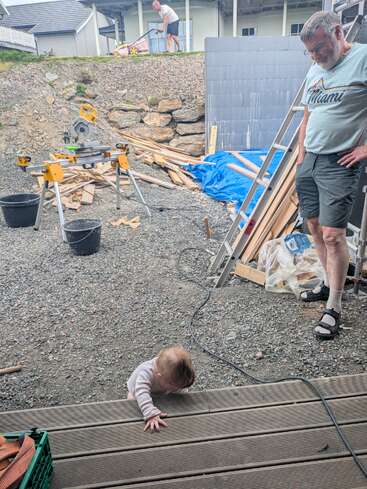 Ein Baby klettert die Holzstufen hinauf, während ein älterer Mann in der Nähe zusieht. Baumaterialien, Werkzeuge und Holz liegen auf dem Hof verstreut. Eine andere Person mäht oben.