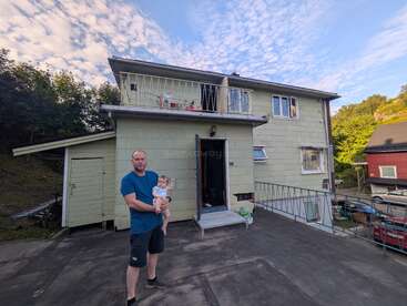 Ein Mann in einem blauen Hemd hält ein Baby vor einem zweistöckigen, hellgrünen Haus. Der Himmel ist blau mit vereinzelten Wolken. Das Haus ist von Bäumen umgeben.