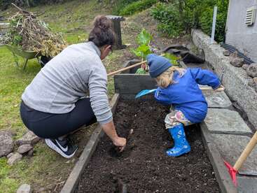 Eine Frau und ein Kind arbeiten zusammen im Garten und graben in der Erde eines Hochbeetes. Das Kind trägt blaue Stiefel und eine Mütze.