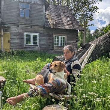 Une femme se détend dans un hamac rustique en compagnie de son chien, entourée de hautes herbes, de fleurs sauvages et d'une vieille maison en bois, par une journée ensoleillée.