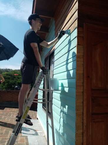 Uma pessoa está em uma escada pintando o exterior de madeira de uma casa de azul claro. Ele usa camisa preta, luvas e óculos, trabalhando sob a luz do sol.