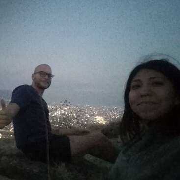 Dos personas están sentadas en la ladera de una colina al atardecer, sonriendo y disfrutando de una vista panorámica de la ciudad con luces centelleantes. Una de las personas levanta el pulgar.