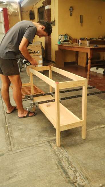 A man is building a wooden table in a workshop area, using clamps and tools. There’s a workbench, some tools, and a dog resting nearby.