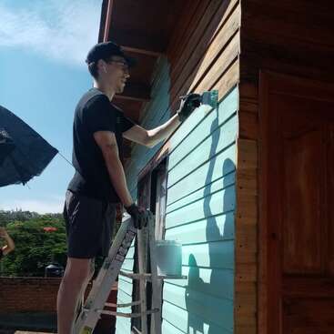 Une personne se tient sur une échelle et peint l'extérieur en bois d'une maison en bleu clair. Elle porte une chemise noire, des gants et des lunettes, et travaille sous la lumière du soleil.