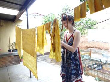 Una mujer con un vestido estampado cuelga ropa amarilla en un tendedero al aire libre. Luz diurna intensa, entorno rústico, plantas y ladrillos al fondo, ambiente desenfadado y creativo.