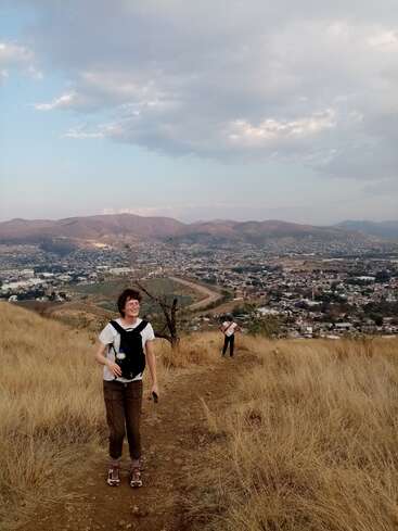 Dos excursionistas suben por un sendero seco y cubierto de hierba desde el que se divisa una extensa localidad enclavada en un valle, rodeada de montañas bajo un cielo parcialmente nublado.