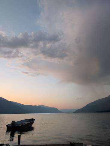 Um pequeno barco flutua em um lago calmo cercado por montanhas ao pôr do sol, com nuvens dramáticas e cores pastéis suaves preenchendo o tranquilo céu noturno.