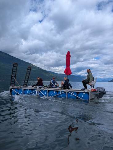 Quatre personnes sont assises sur un radeau artisanal fait de tonneaux et de métal, flottant sur un lac calme, sous un ciel nuageux, avec des montagnes en arrière-plan.