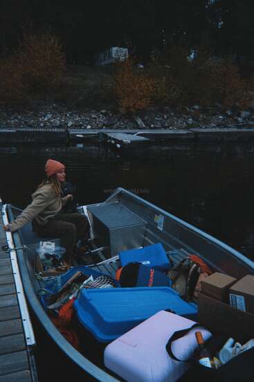Une femme portant un bonnet est assise dans un petit bateau rempli de provisions, amarré au bord d'un lac avec des arbres d'automne et une caravane en arrière-plan.