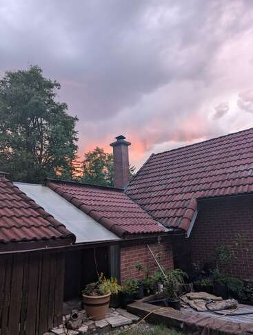Une maison en briques avec des toits en tuiles rouges se trouve sous un ciel nuageux au coucher du soleil, entourée d'arbres, de plantes, de pots de fleurs, d'outils de jardinage et de matériaux d'extérieur.