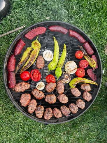 Uma churrasqueira redonda cozinhando hambúrgueres de carne, salsichas fatiadas, pimentões verdes, tomates e cebolas. A churrasqueira é colocada ao ar livre sobre a grama verde, pronta para um banquete.