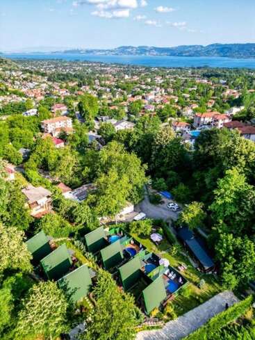 Essa imagem aérea captura uma cidade verde e exuberante com grupos de casas, cabanas com telhados verdes, estradas sinuosas e um lago panorâmico emoldurado por montanhas distantes.
