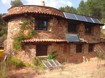 A imagem mostra uma casa de pedra rústica com painéis solares no telhado, cercada por árvores e um ambiente natural sereno.
