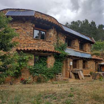 La imagen muestra una casa rústica de piedra con paneles solares en el tejado, rodeada de exuberante vegetación y un cielo nublado, que destila una atmósfera natural y serena.