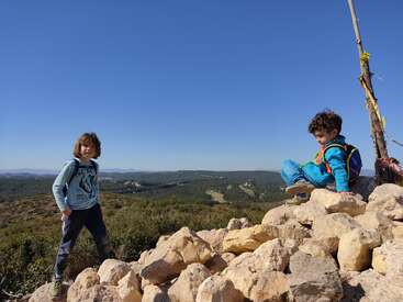 Two children stand on a rocky hilltop, with one sitting atop a pile of rocks and the other standing beside them, set against a clear blue sky.