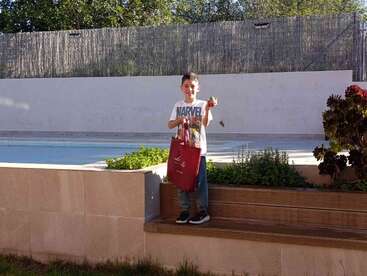 A young boy stands beside a swimming pool, holding a red bag and a small green object, dressed in a Marvel shirt, surrounded by plants and sunlight.