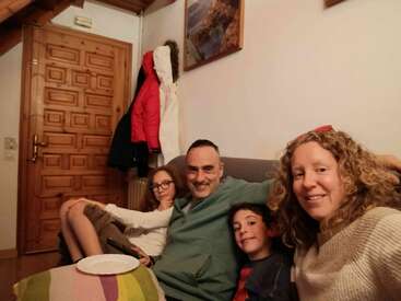 A happy family of four sits closely together on a couch, smiling at the camera in a cozy living room with wooden door, coats, and framed pictures.