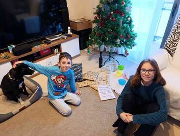 Two children sit near a decorated Christmas tree with wrapped presents, a dog in a bed, and holiday treats on the floor, creating a festive atmosphere.