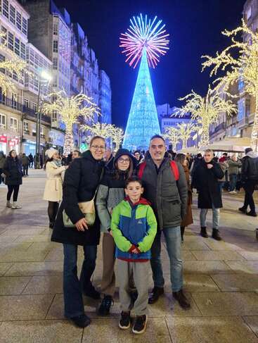 A family poses in front of a large, illuminated Christmas tree at night, surrounded by festive lights and people in a lively city street.
