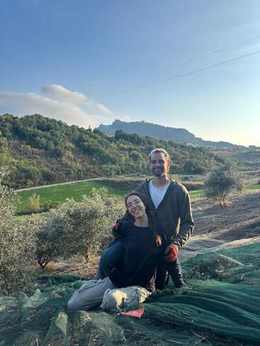 Una pareja sonriente posa al aire libre sobre una red verde en un paisaje pintoresco, rodeada de olivos y colinas, con un cielo azul despejado sobre ellos.