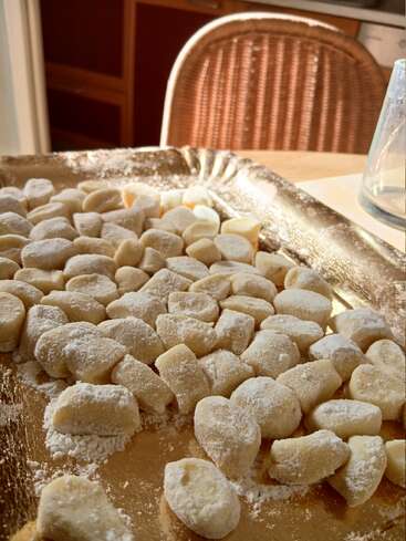 Una bandeja dorada contiene ñoquis recién hechos, crudos y espolvoreados con harina, sobre una mesa en una cocina iluminada por el sol, listos para ser cocinados.