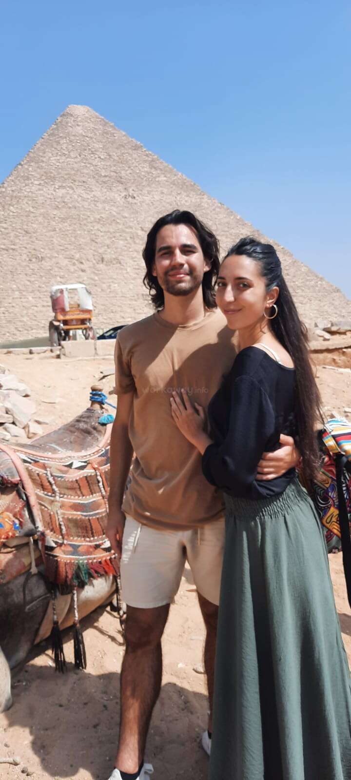 Una pareja posa feliz frente a la Gran Pirámide de Egipto, con un burro cerca bajo el cielo azul brillante, creando un recuerdo de viaje.
