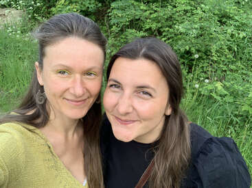 Dos mujeres sonríen y posan para hacerse un selfi al aire libre. Están muy juntas, rodeadas de exuberante vegetación y césped, y parecen felices y satisfechas.