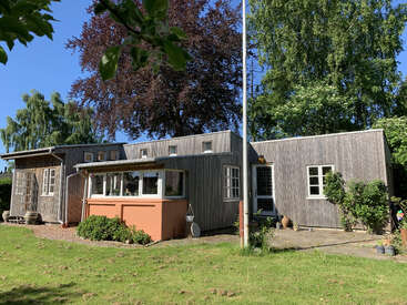 This image shows a rustic wooden house with a small porch, surrounded by green grass, large trees, and clear blue skies on a sunny day.