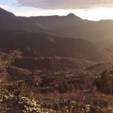 The image depicts a serene mountainous landscape with lush vegetation, featuring a small village nestled among the hills, bathed in warm sunlight.