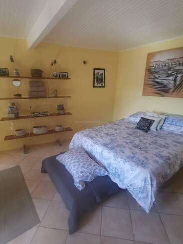 Cette chambre confortable comprend un lit double avec une literie à motifs, des murs jaunes, des étagères en bois ouvertes avec des objets de décoration, un tableau nautique, un sol carrelé et un éclairage doux et accueillant.