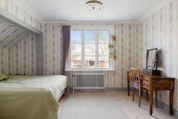 Une chambre à coucher confortable et vintage avec un papier peint à motifs, un lit avec une couverture verte, un meuble-lavabo en bois, une fenêtre, un radiateur, des plantes et une lumière naturelle intense.