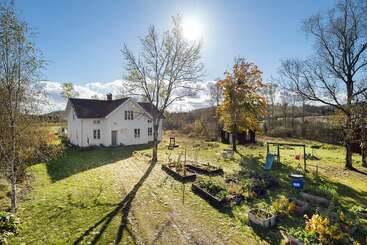 Une charmante maison blanche se dresse sur un terrain ensoleillé et herbeux, avec une aire de jeux, des parterres de jardin, des arbres et un magnifique paysage rural sous un ciel bleu éclatant.
