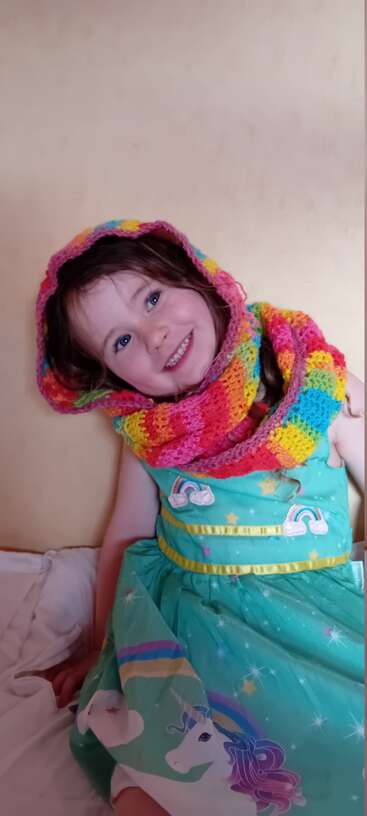 A smiling young girl in a rainbow crocheted hooded scarf and a teal unicorn dress, sitting happily against a plain wall, radiating joy and warmth.