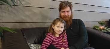 A bearded man and a young girl sit closely together on a black couch. The girl wears a pink striped shirt, and both appear happy and relaxed.