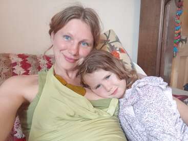 A smiling woman and a young child cuddle on a couch. The woman wears a green top and the child has a floral dress. Cozy, loving atmosphere.