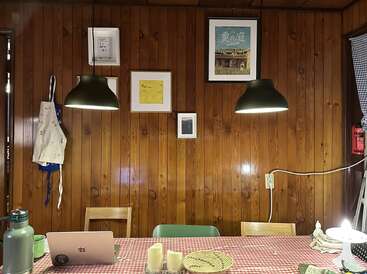 A cozy dining area with wooden walls, two hanging lamps, framed pictures, an apron, laptop, mugs, candles, a basket, and a checkered red tablecloth.