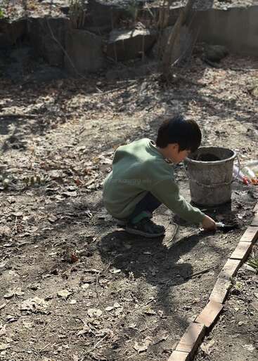 A young child is squatting on bare ground, digging with a small tool next to a bucket, surrounded by dry leaves and sunlight in a garden.