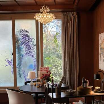 A cozy room features a round table with flowers, lamp, snacks, and a book. Large windows display colorful doodles. Warm wood tones, chandelier, and map decorate.