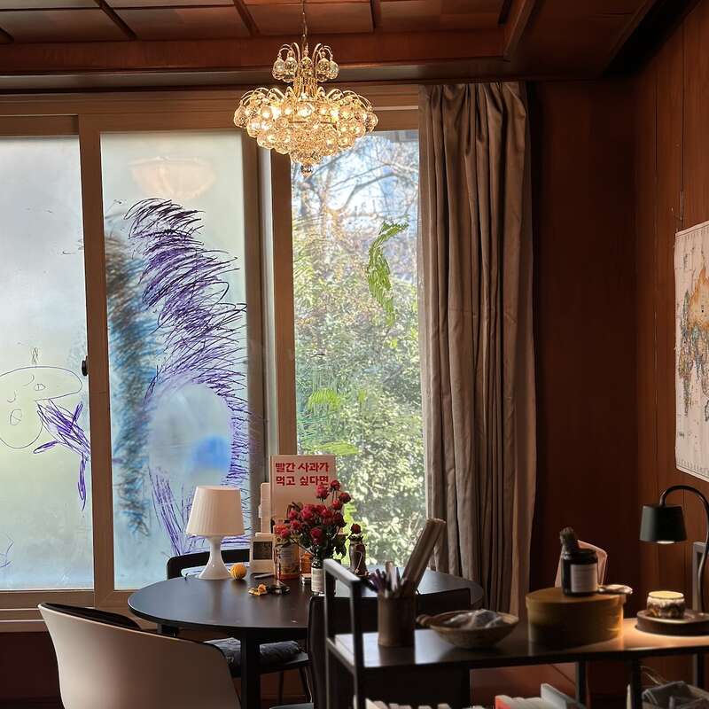 A cozy room features a round table with flowers, lamp, snacks, and a book. Large windows display colorful doodles. Warm wood tones, chandelier, and map decorate.