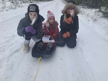 Tres niños, abrigados con trajes de invierno, posan en una carretera nevada con un trineo y bastones de caramelo, rodeados de un sereno paisaje invernal.