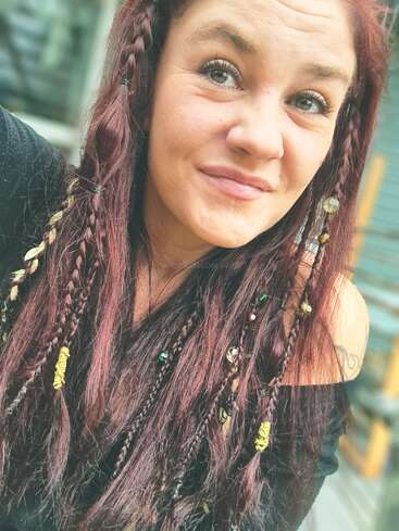 A woman with long reddish-brown hair and braids adorned with beads and string smiles at the camera, wearing a black off-shoulder top outdoors.